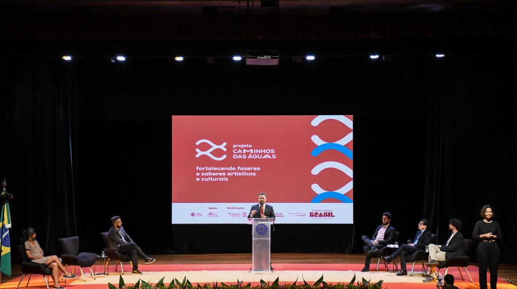 Homem branco de terno e gravata fala no púlpito no centro do palco da universidade. Ao fundo uma projeção da logomarca do projeto Caminhos das Águas em vermelho branco e azul